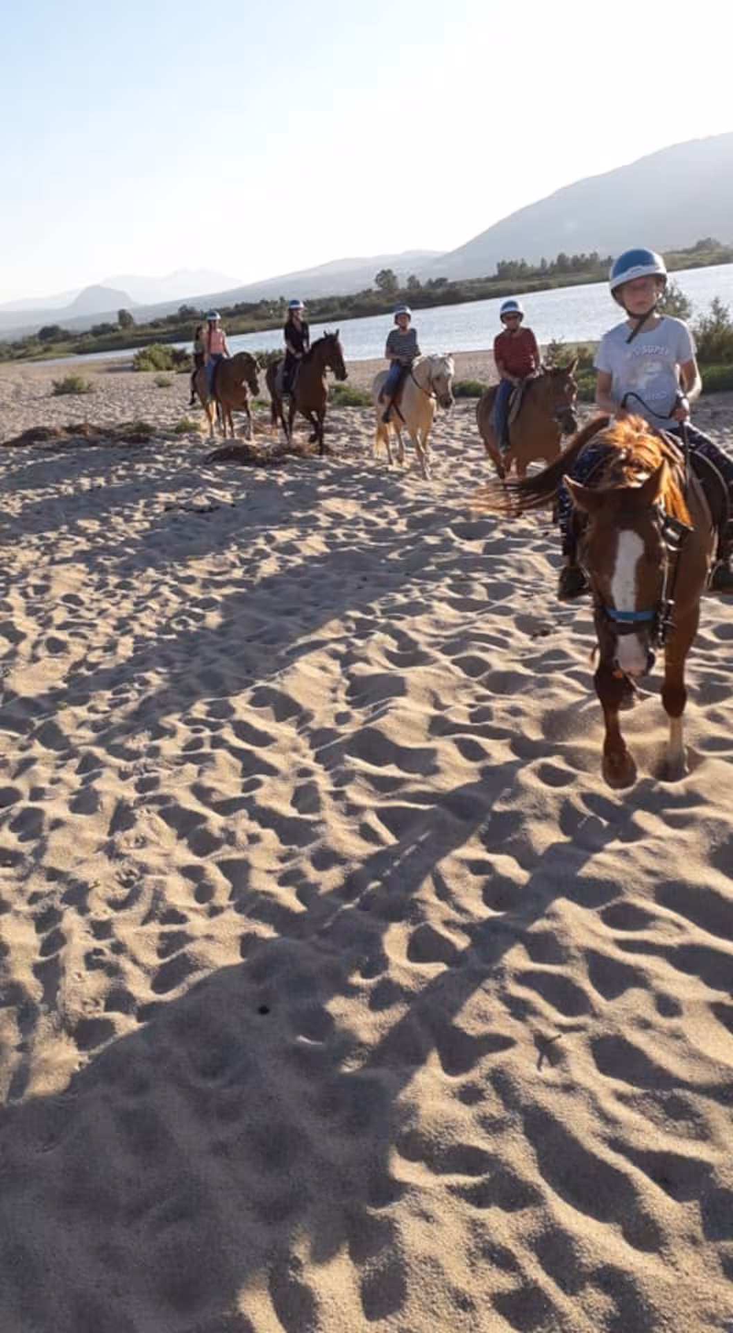 Riders on horseback along the sandy Orosei beach at sunrise, with a serene river and mountains nearby.