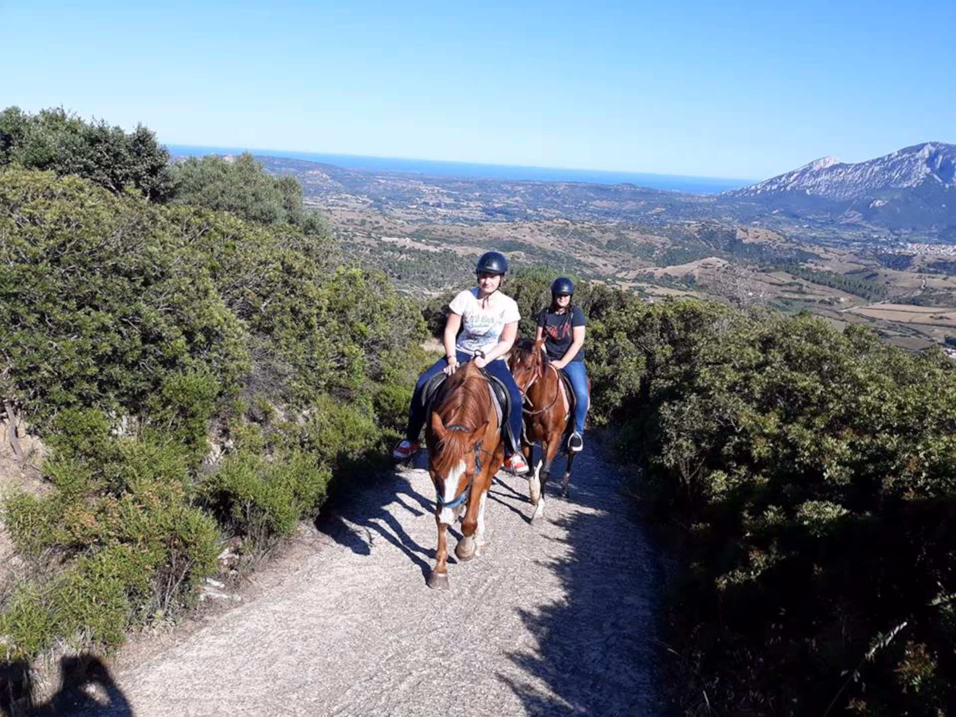 Riders on horseback enjoy panoramic views of Orosei's landscape under a clear blue sky.