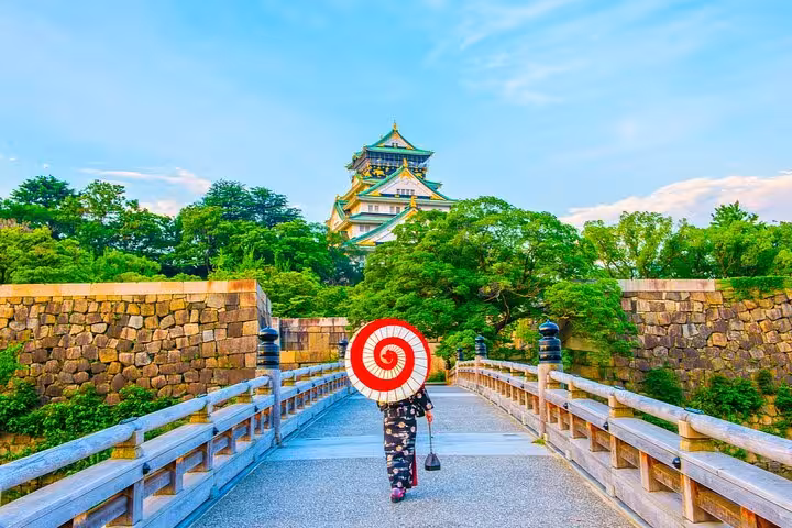 Woman in kimono with red spiral umbrella walking toward Osaka Castle on a historic bridge