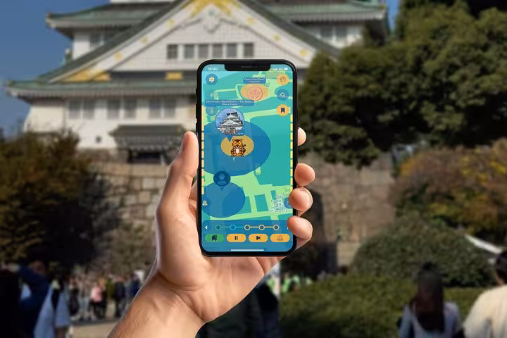 Hand holding smartphone with self-guided tour app in front of Osaka Castle, Japan
