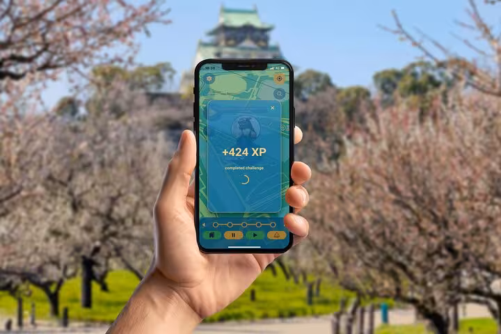 Hand holding smartphone showing Pokémon GO +424 XP with Osaka Castle and cherry blossom trees in background