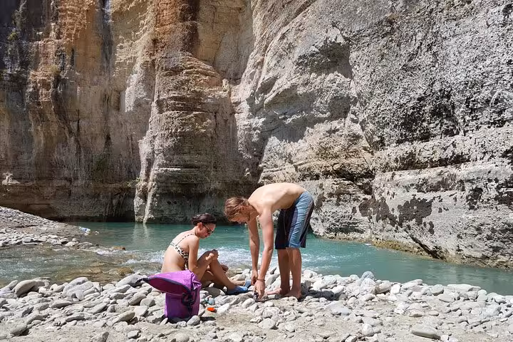 Two people relax by the turquoise waters and rugged cliffs of Osum Canyon, perfect for a nature retreat.