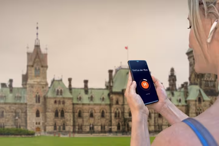 Woman using self-guided audio tour app on smartphone in front of East Block Parliament Hill Ottawa