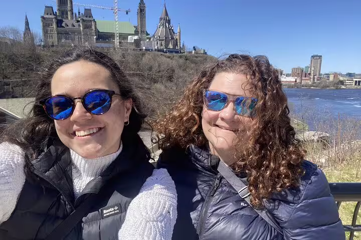 Two smiling women in sunglasses on Ottawa self-guided walking tour with Parliament Hill and Ottawa River behind them