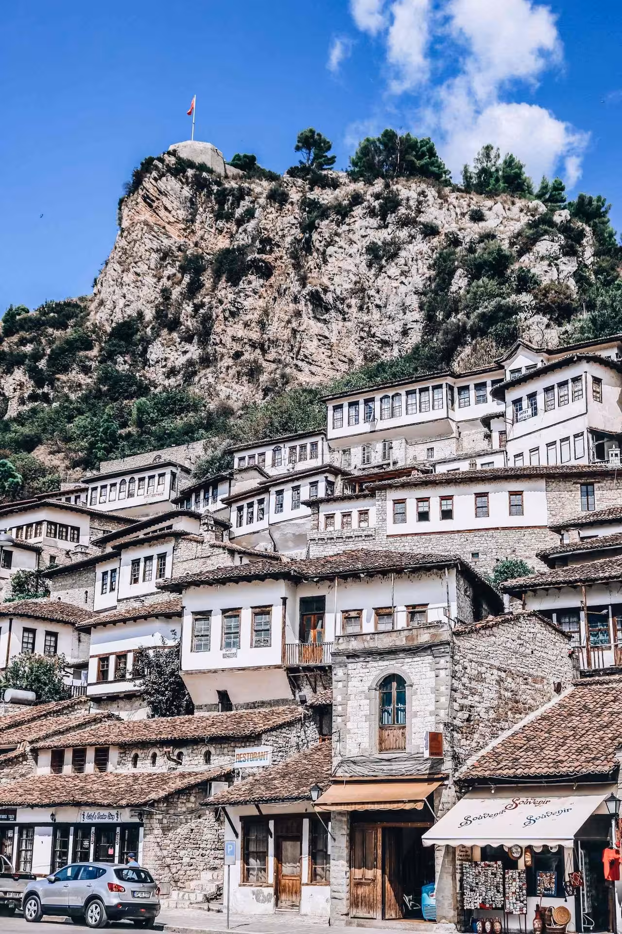 Traditional Ottoman houses in Berat, Albania, set against a rugged hilltop with a flag, showcasing rich cultural heritage.