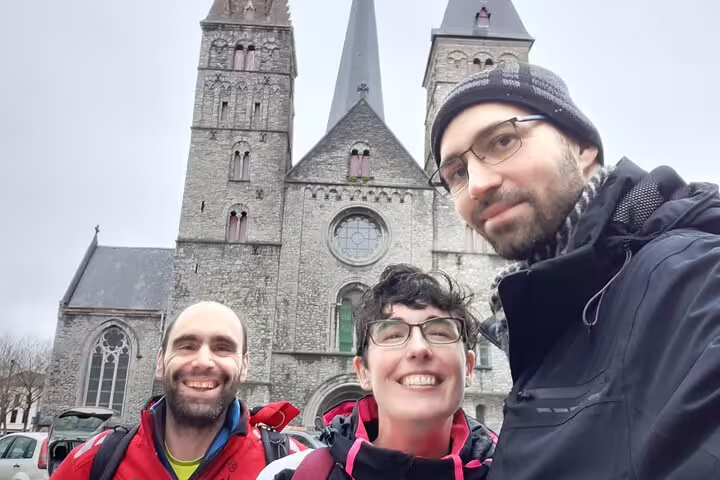 Three smiling hikers with backpacks posing in selfie in front of historic Gothic church in Ghent city center