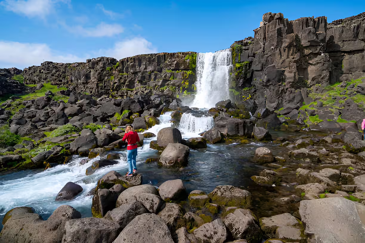 Traveler standing on rocks beside Öxará river with Öxarárfoss waterfall in Iceland's Þingvellir National Park