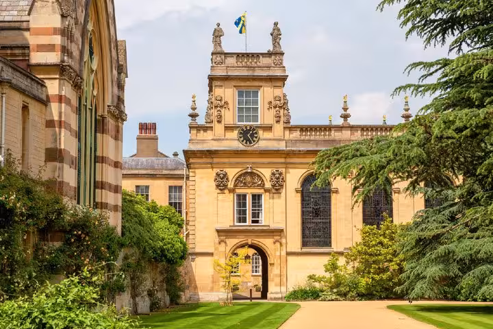 Historic Oxford college gateway with ornate stonework, clock tower, and lush green gardens on a sunny day
