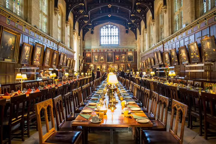 Historic dining hall at Oxford University set for a meal, featuring long wooden tables, ornate chairs, and grand paintings.