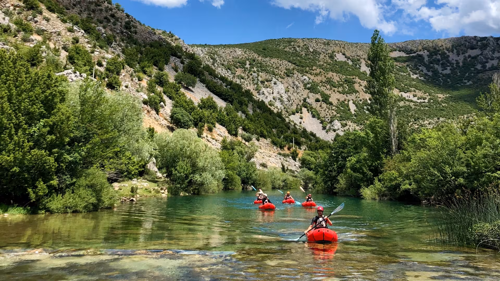 Packrafters enjoying a scenic paddle along the serene Zrmanja River, framed by lush green hills and clear skies.