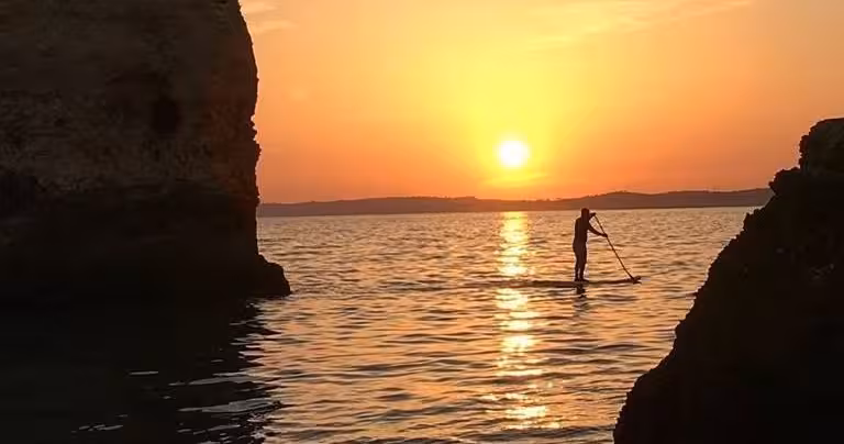 Silhouette of a paddleboarder gliding past rocky cliffs during a serene private sunset boat tour on calm coastal waters