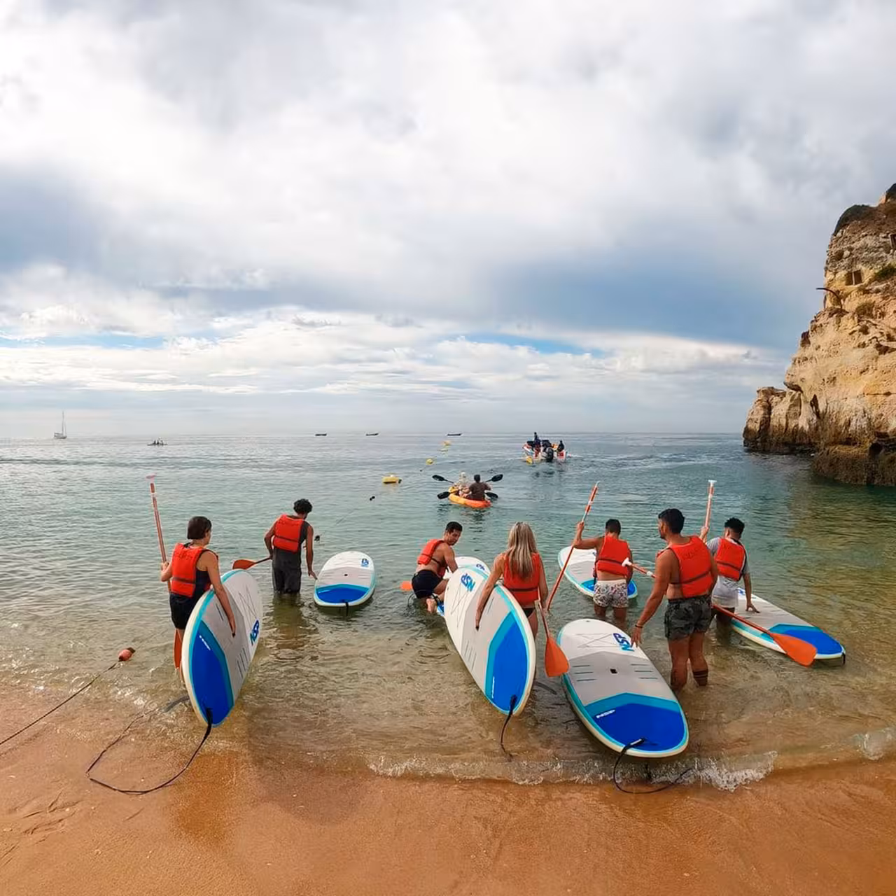 Group of paddleboarders in red vests preparing to enter calm sea waters from a sandy beach, perfect for beginners.
