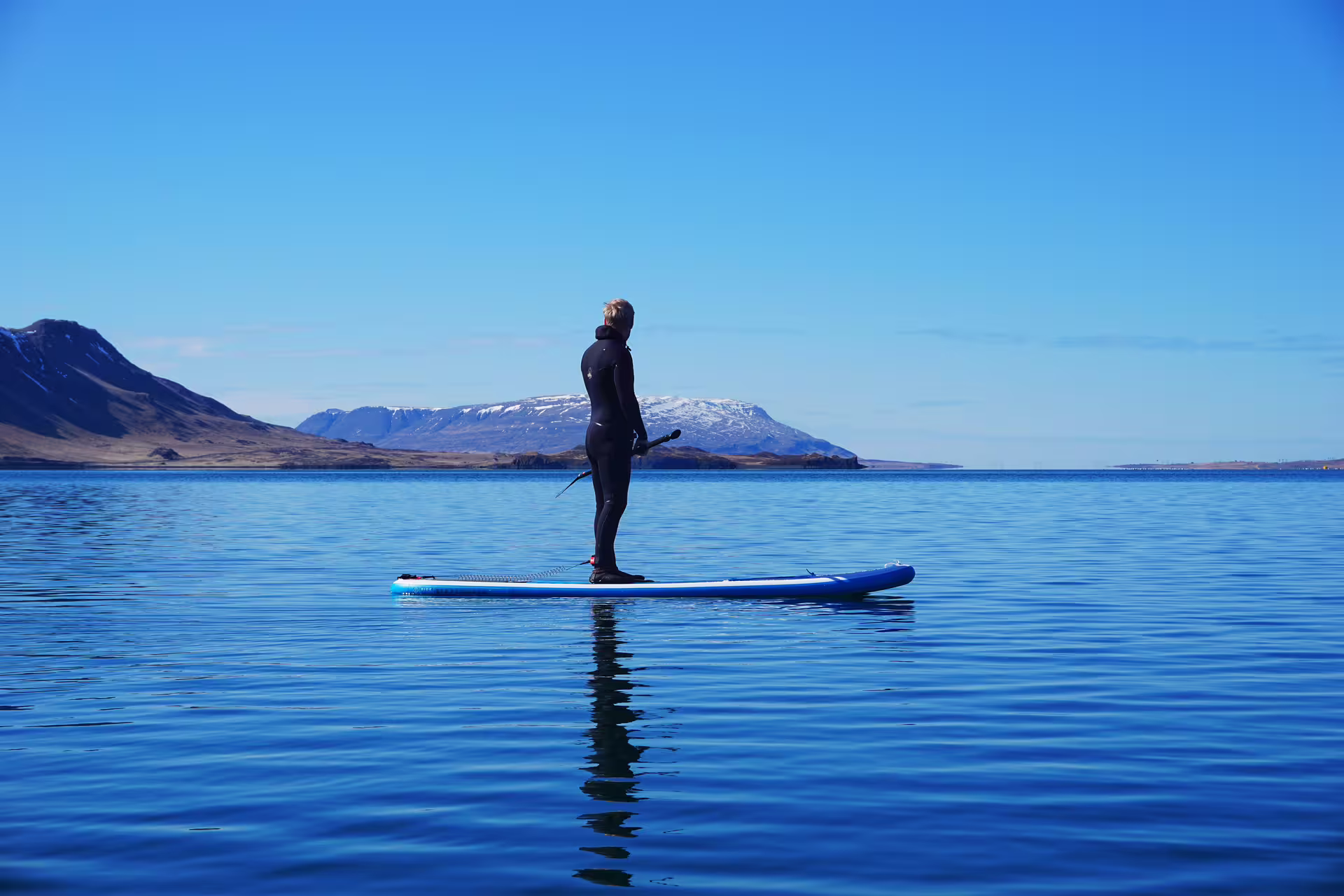 Person paddleboarding in serene, crystal-clear waters of the Forgotten Fjords, surrounded by majestic snow-capped mountains.