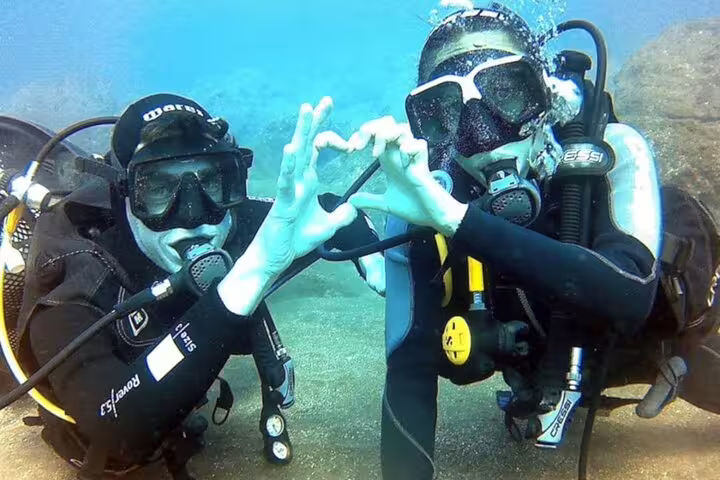 Two scuba divers in wetsuits make a heart shape with their hands underwater during an open water diving course adventure.