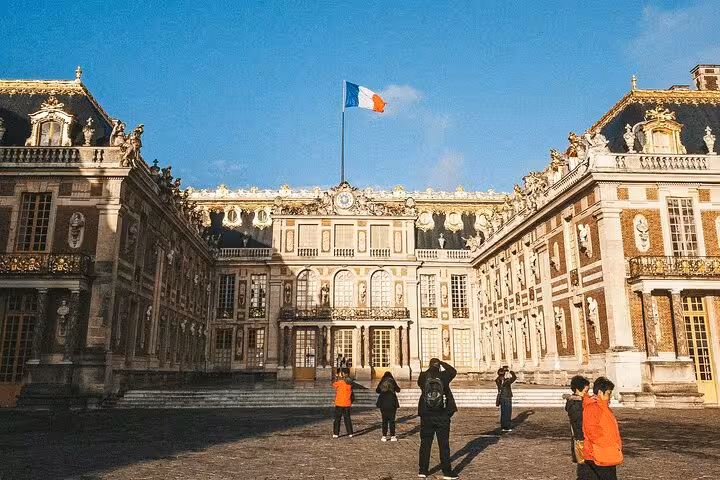 Tourists visiting the ornate facade of the Palace of Versailles with French flag flying above courtyard