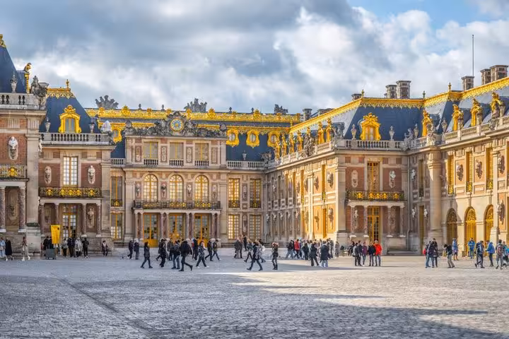Tourists walking in the marble courtyard of the Palace of Versailles with ornate golden facades