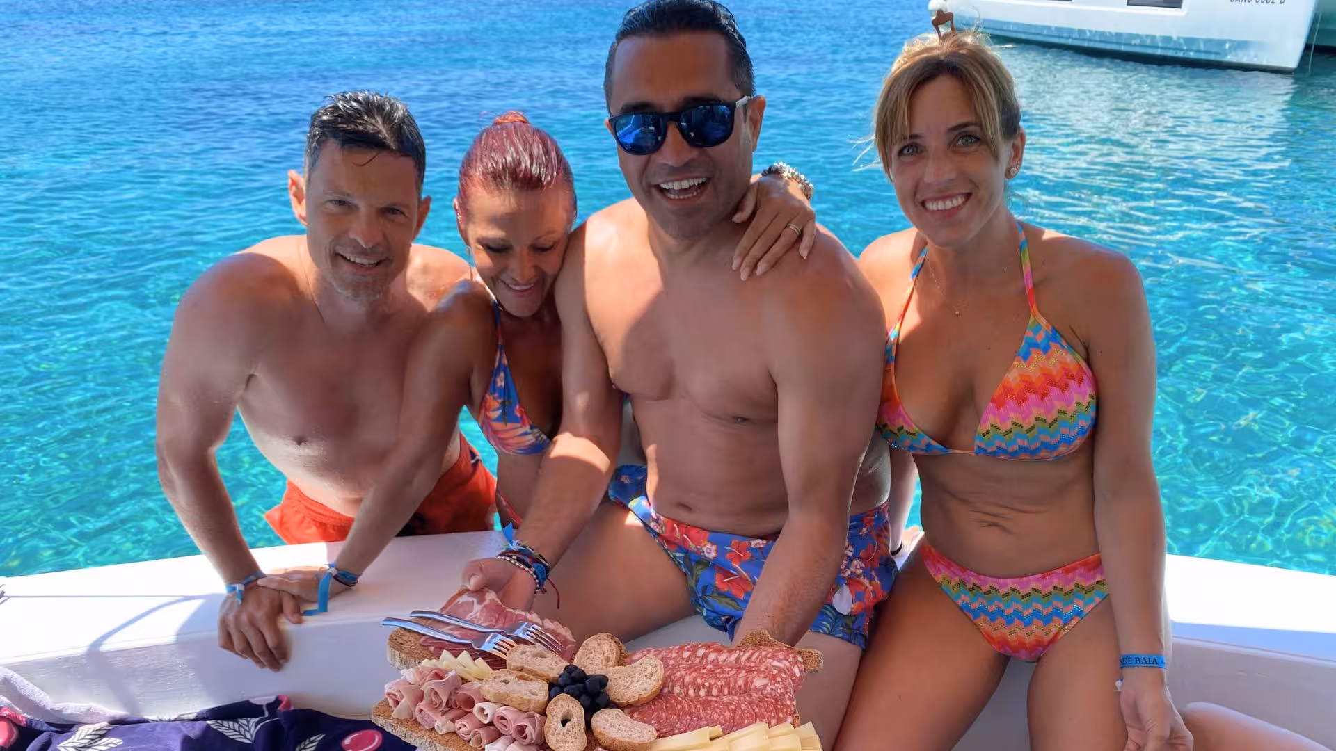 Group enjoying a delicious charcuterie platter on a boat during a Palau or La Maddalena tour with turquoise waters.