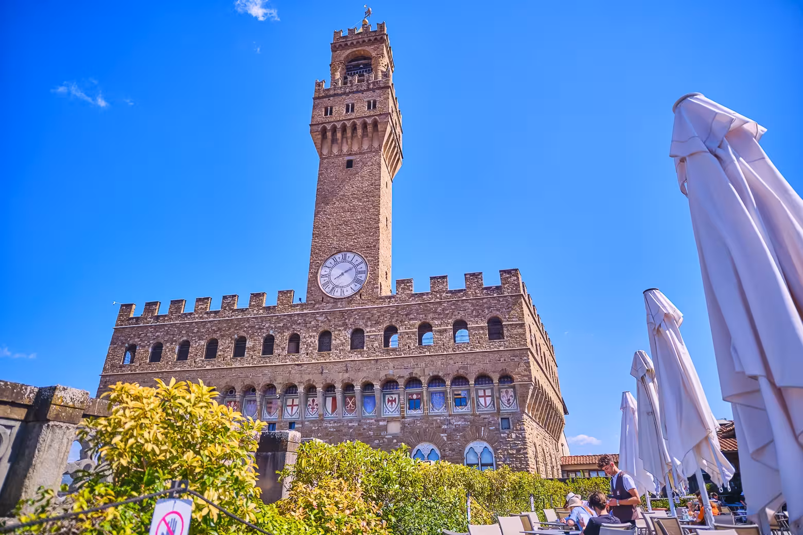 View of Palazzo Vecchio's clock tower under a blue sky, seen from a nearby terrace in Florence, Italy.