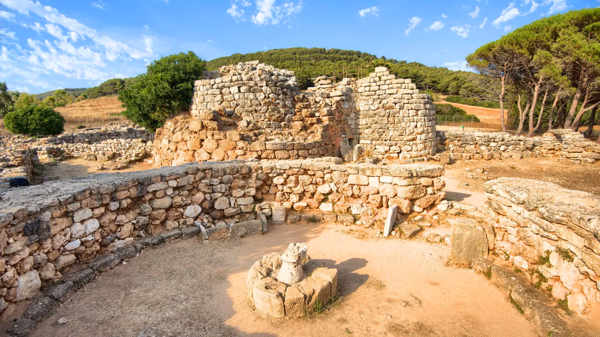 Explore the ancient stone structures of Palmavera nuraghe in Alghero, surrounded by lush greenery and clear skies.