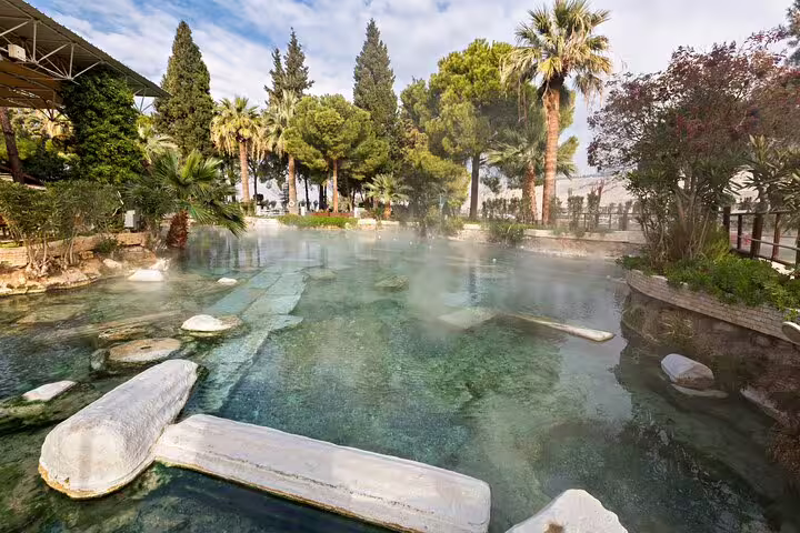 Ancient thermal pool at Pamukkale surrounded by lush greenery and historic ruins, offering a serene natural retreat.