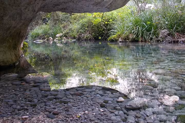 Serene view of a clear stream under a rocky overhang surrounded by lush greenery in Pantalica, Sicily.