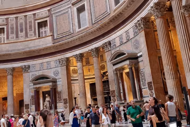 Interior of the Pantheon in Rome, featuring ornate columns, detailed frescoes, and a crowd of visitors exploring.