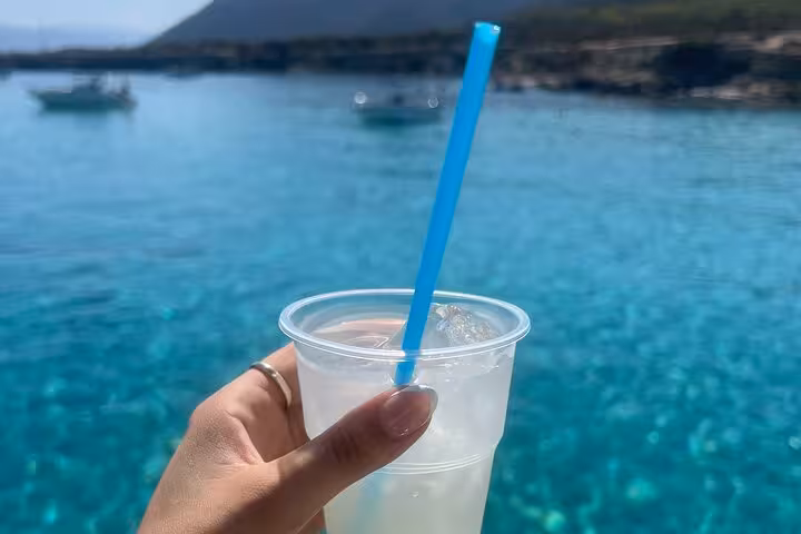 Hand holding cold drink on boat with Akamas Blue Lagoon turquoise sea in background on Paphos day trip