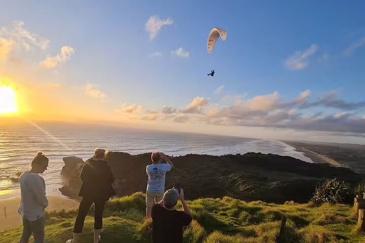 Paraglider soaring over Auckland coastline at sunset, capturing stunning views on a romantic tour with winery and thermal pool.