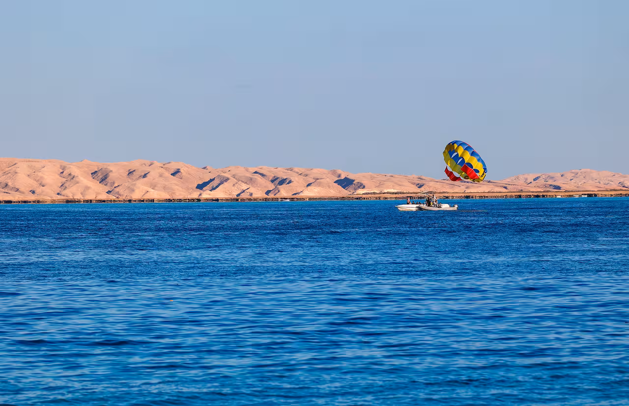Parasailing trip boat towing a bright parachute over calm blue sea with desert coastline in the distance