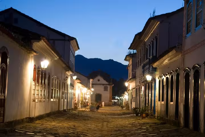Evening view of Paraty's illuminated colonial buildings along cobblestone streets with mountain silhouette in the background.