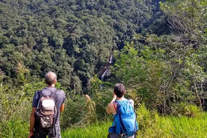 Two hikers with backpacks admire the distant Melancia Waterfall surrounded by dense forest on a vibrant Paraty tour.