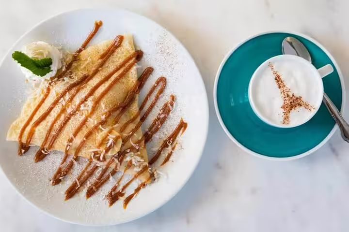 Caramel drizzled crepe with powdered sugar and a cappuccino with Eiffel Tower latte art on teal saucer