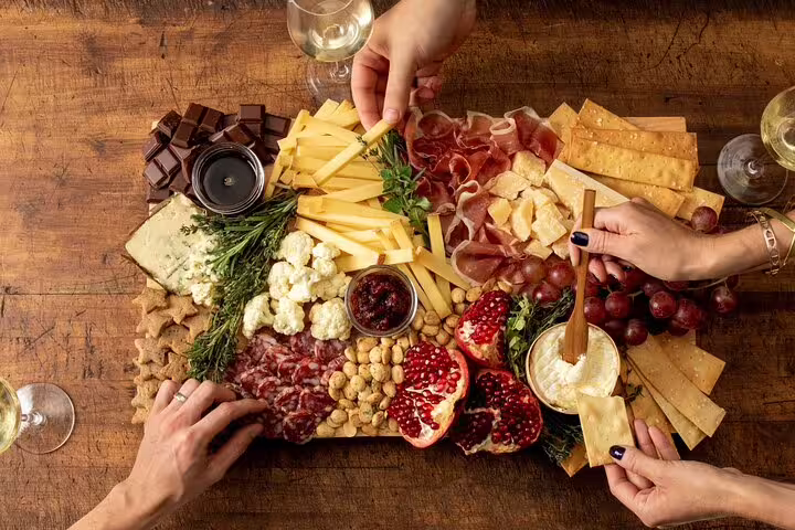 Hands reaching for a charcuterie board with cheese, cured meats, pomegranate, nuts, crackers and wine glasses