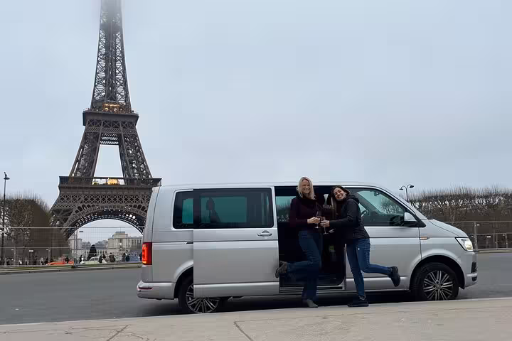 Two women toasting with champagne beside a silver minivan with the Eiffel Tower in Paris behind them.