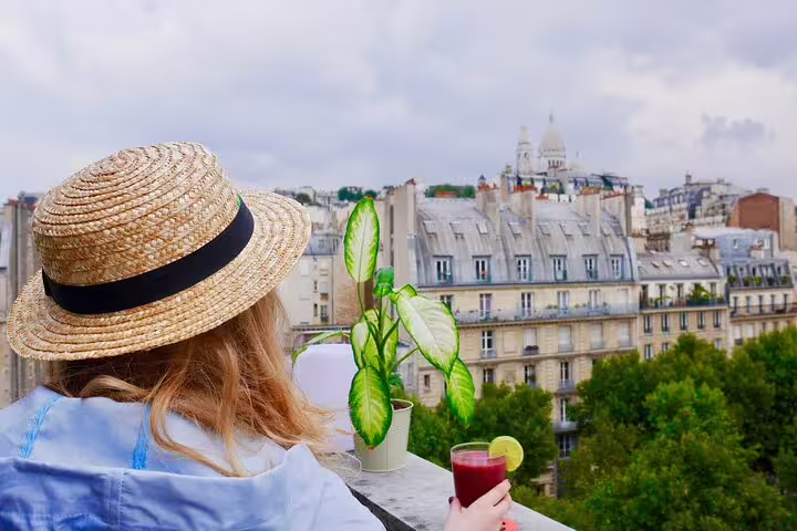 Woman in straw hat enjoying a drink on a Paris rooftop with Sacré-Cœur and Montmartre rooftops in view