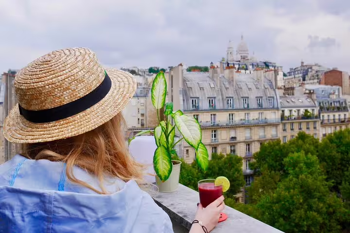 Woman in straw hat holding red drink on rooftop terrace with Sacré-Cœur and Montmartre rooftops in background