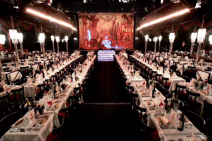 Elegant Parisian cabaret dining hall with long white-clothed tables, candles, and ornate red stage backdrop