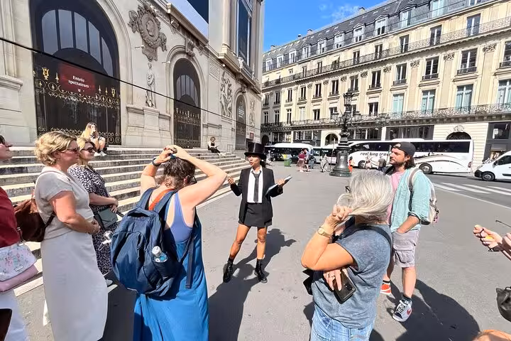 Tour guide in black hat and suit leads group of tourists outside Paris Opera House on sunny day
