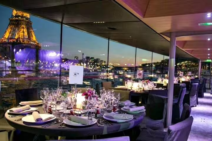 Elegant dinner cruise table setting with Eiffel Tower illuminated at night through panoramic windows on Seine River