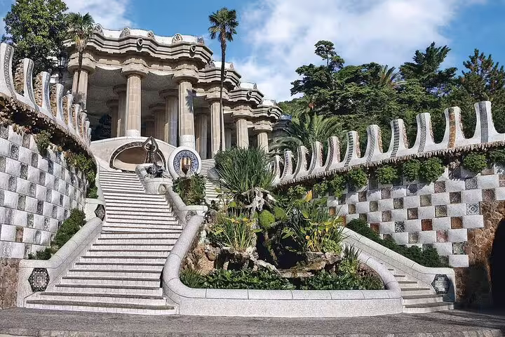 Ornate staircase and columned pavilion at Park Güell, Barcelona, with mosaic railings and lush greenery