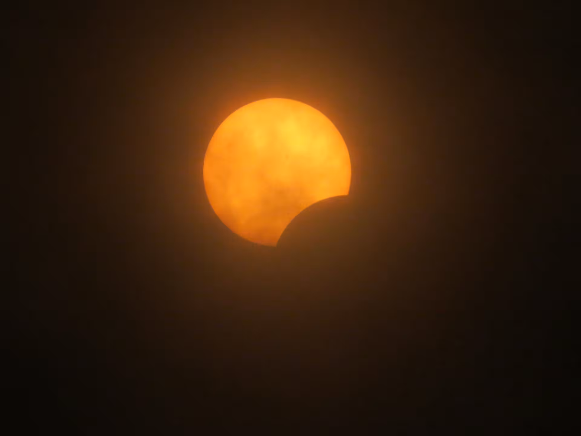 Partial solar eclipse with the moon covering lower right of the orange sun through hazy clouds
