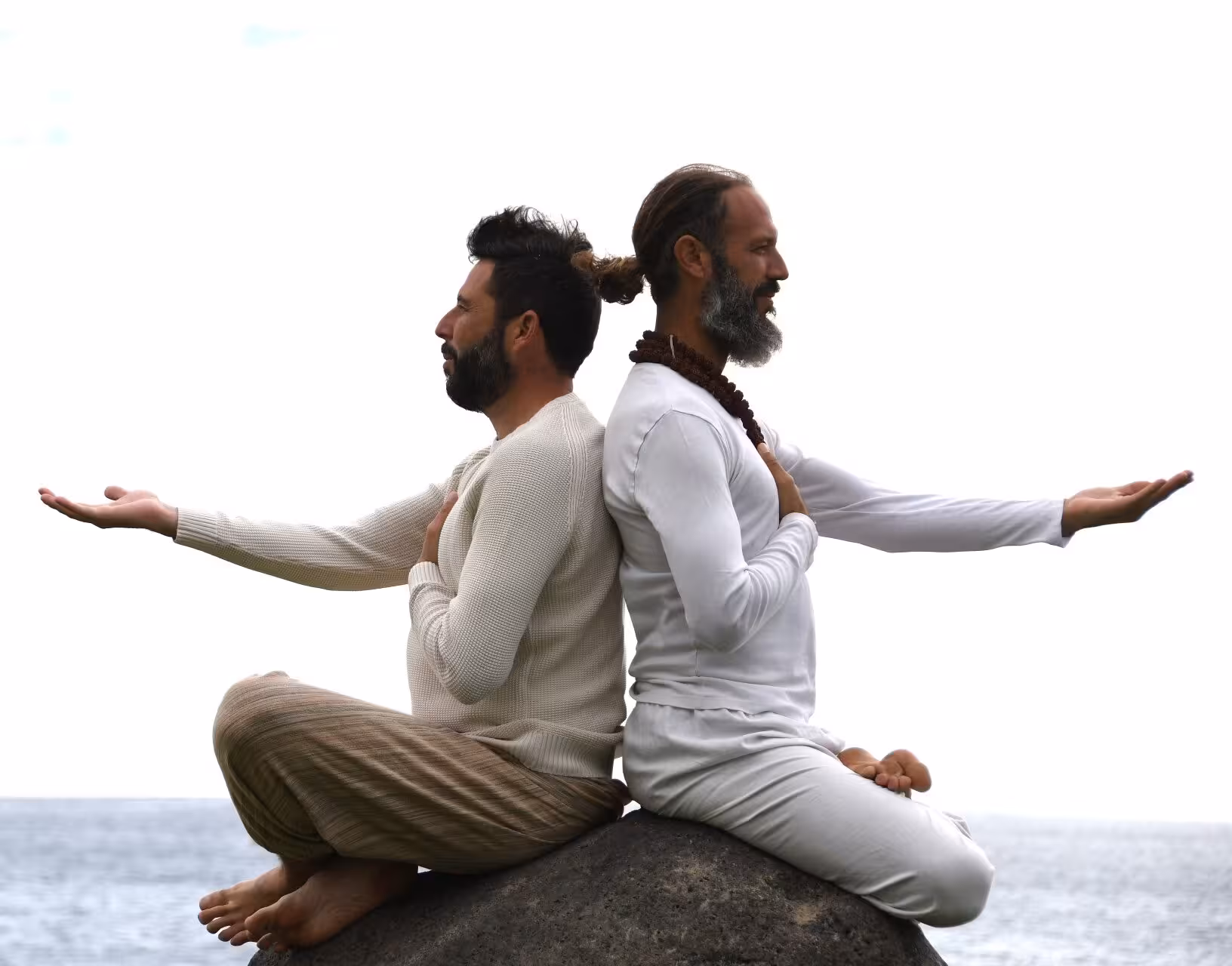 Two men in white seated back-to-back in lotus pose with arms outstretched on a rock by the ocean