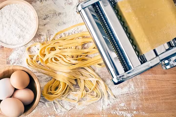 Close-up of a pasta machine with fresh dough, eggs, and flour on a wooden surface in a cooking class.
