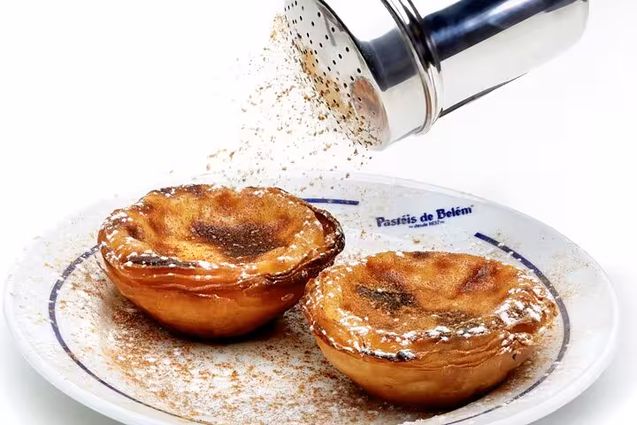 Close-up of traditional Pastéis de Belém being dusted with cinnamon and sugar on a white plate.