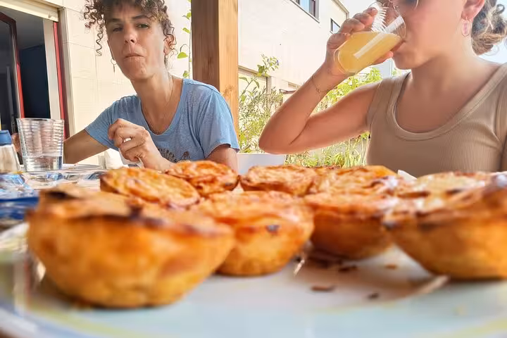 Two people enjoy Pastel De Nata and drinks on a sunny terrace, highlighting a relaxed Portuguese dining experience.