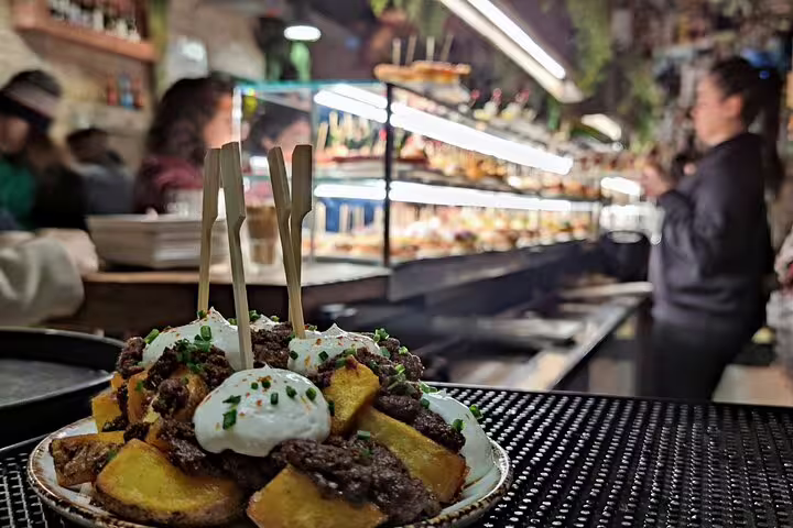 Plate of patatas bravas topped with aioli and minced meat in a bustling Barcelona tapas bar.