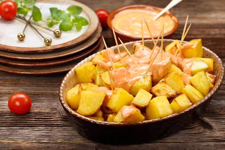 A rustic dish of patatas bravas with spicy sauce, served in a traditional Barcelona tavern setting with cherry tomatoes and herbs.