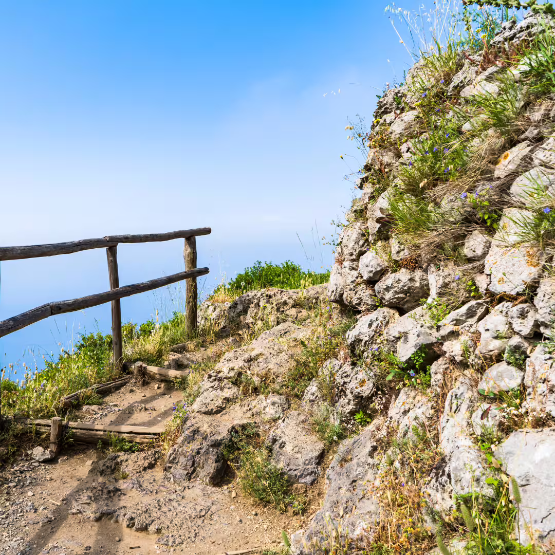 Rocky section of the Path of the Gods hiking trail with wildflowers, rustic fence and panoramic Amalfi Coast sea views