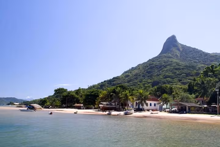 Tranquil beach with boats and lush greenery at the base of Peak Sugar Bread, under a clear blue sky.