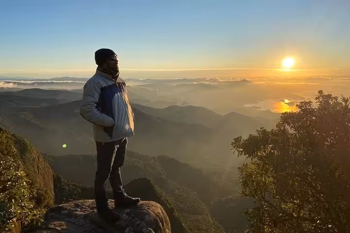 Person stands on Pedra da Macela at sunrise, enjoying panoramic views of misty mountains and vibrant skies.